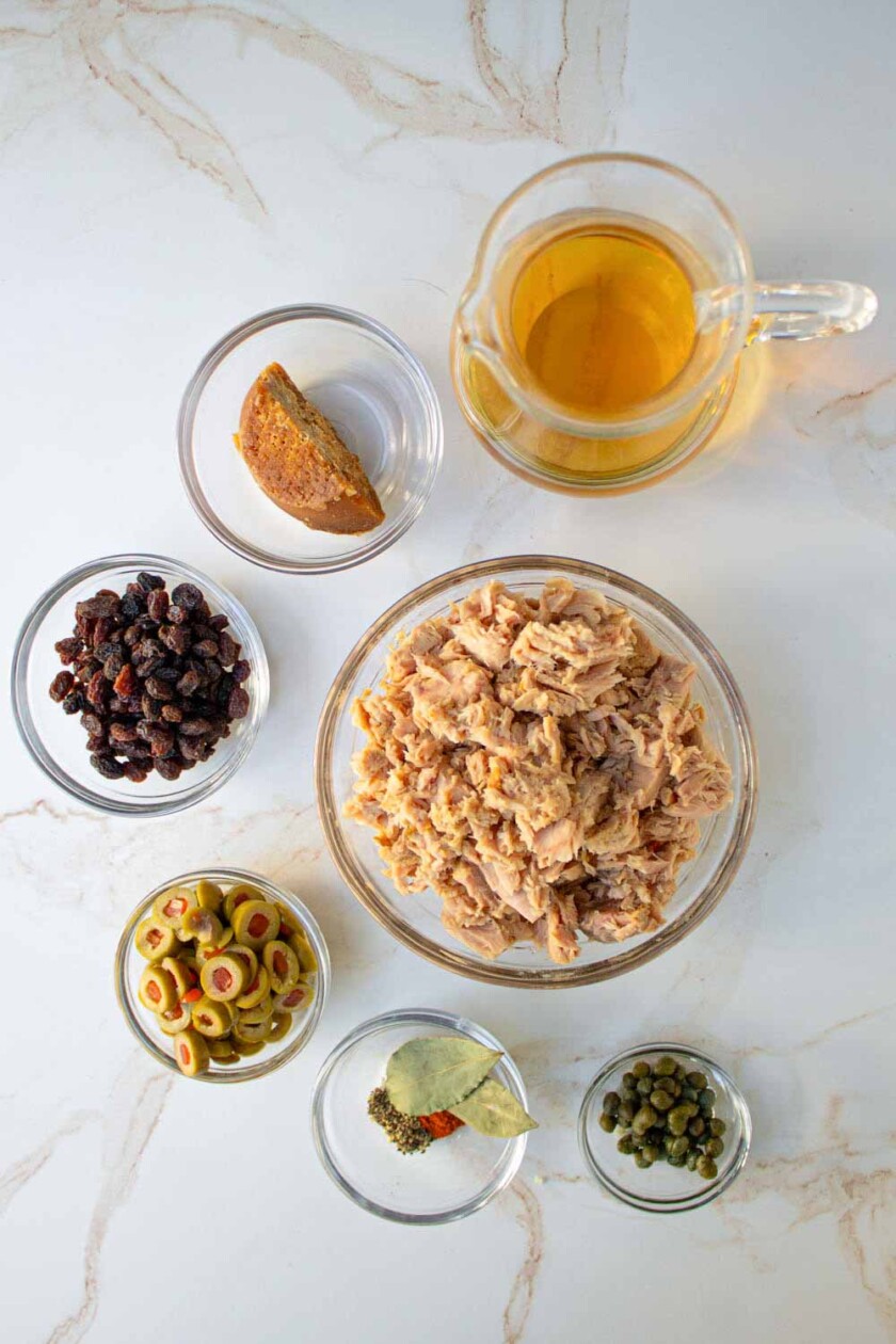Vista aérea de una encimera de cocina con ingredientes para cocinar: atún en un bol, aceitunas, alcaparras, pasas, condimentos y una jarra de vino de cocina.