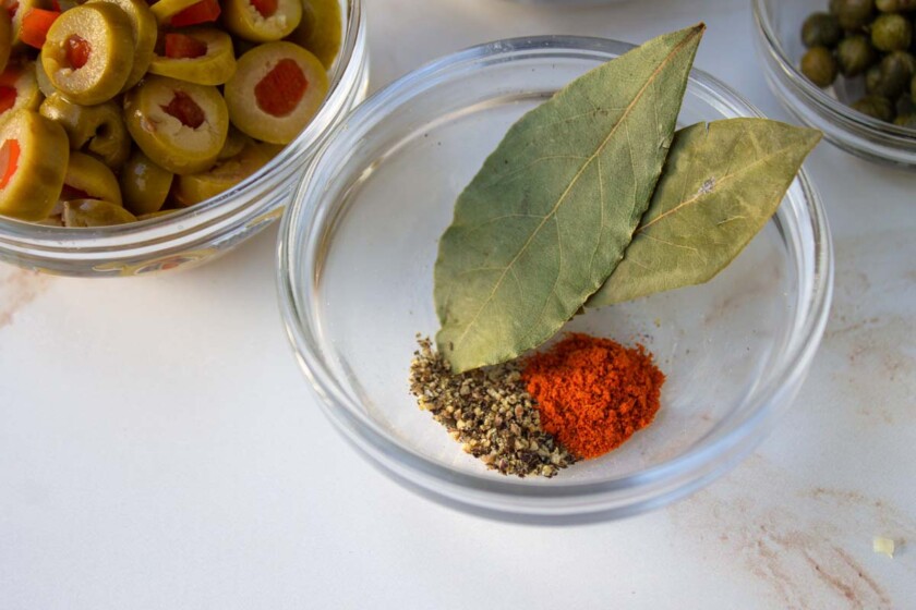 Cuencos de vidrio con especias como pimentón y pimienta negra, pimienta cayena. hojas de laurel y aceitunas verdes en rodajas rellenas de pimientos sobre una superficie blanca.