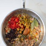 Una sartén de acero inoxidable que contiene una mezcla de tomates picados, aceitunas, alcaparras, pasas y atún, sazonados con hojas de laurel y hojuelas de chile, sobre una superficie de mármol.