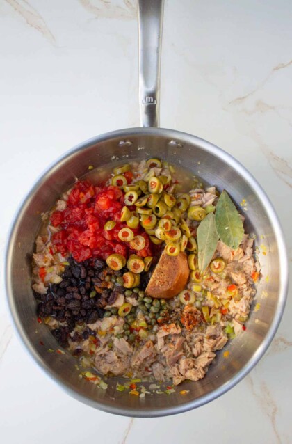 Una sartén de acero inoxidable que contiene una mezcla de tomates picados, aceitunas, alcaparras, pasas y atún, sazonados con hojas de laurel y hojuelas de chile, sobre una superficie de mármol.