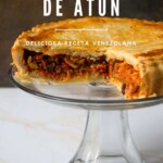 Un pastel de polvorosa de atún cortado sobre un soporte, con texto superpuesto que lo describe como una deliciosa receta venezolana.