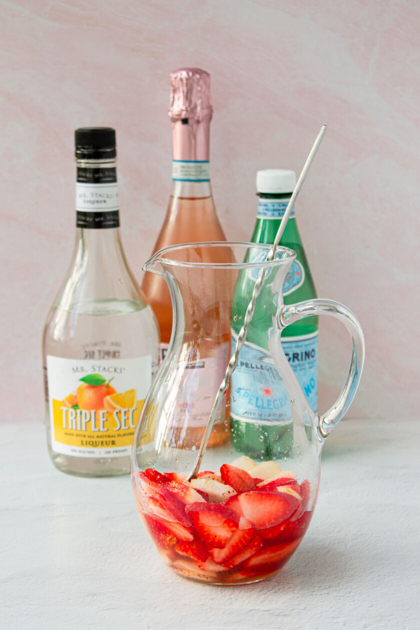 Una selección de ingredientes para un cóctel que incluye fresas frescas, una botella de triple sec, vino espumoso y jarabe de limón.
