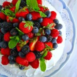 Una pavlova cubierta con una variedad de frambuesas frescas, moras, arándanos y hojas de menta en un plato decorativo de encaje blanco encima de un plato con borde azul.