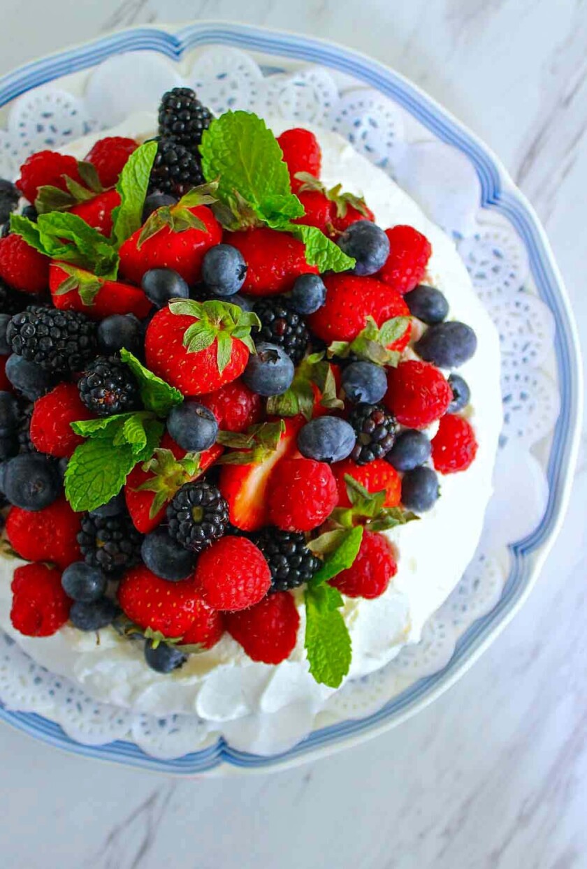 Una pavlova cubierta con una variedad de frambuesas frescas, moras, arándanos y hojas de menta en un plato decorativo de encaje blanco encima de un plato con borde azul.