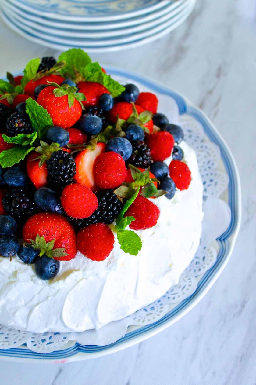 A pavlova dessert topped with an assortment of fresh berries and mint leaves, presented on a decorative plate with a lace-like doily pattern.