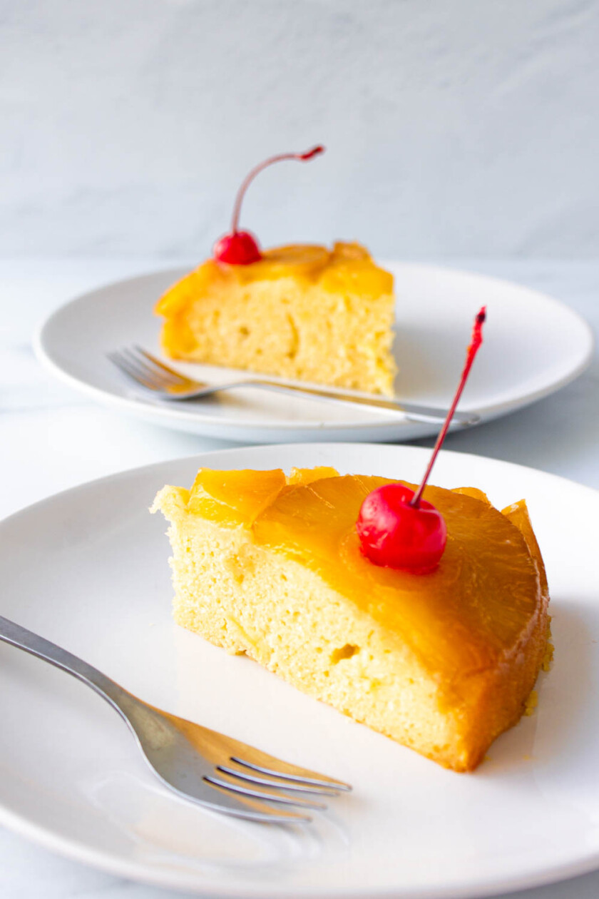 Torta-de-piña-volteada-by-enrilemoine-14 Rebanadas de pastel de piña volteado con cerezas sobre platos blancos con tenedores sobre un fondo claro.