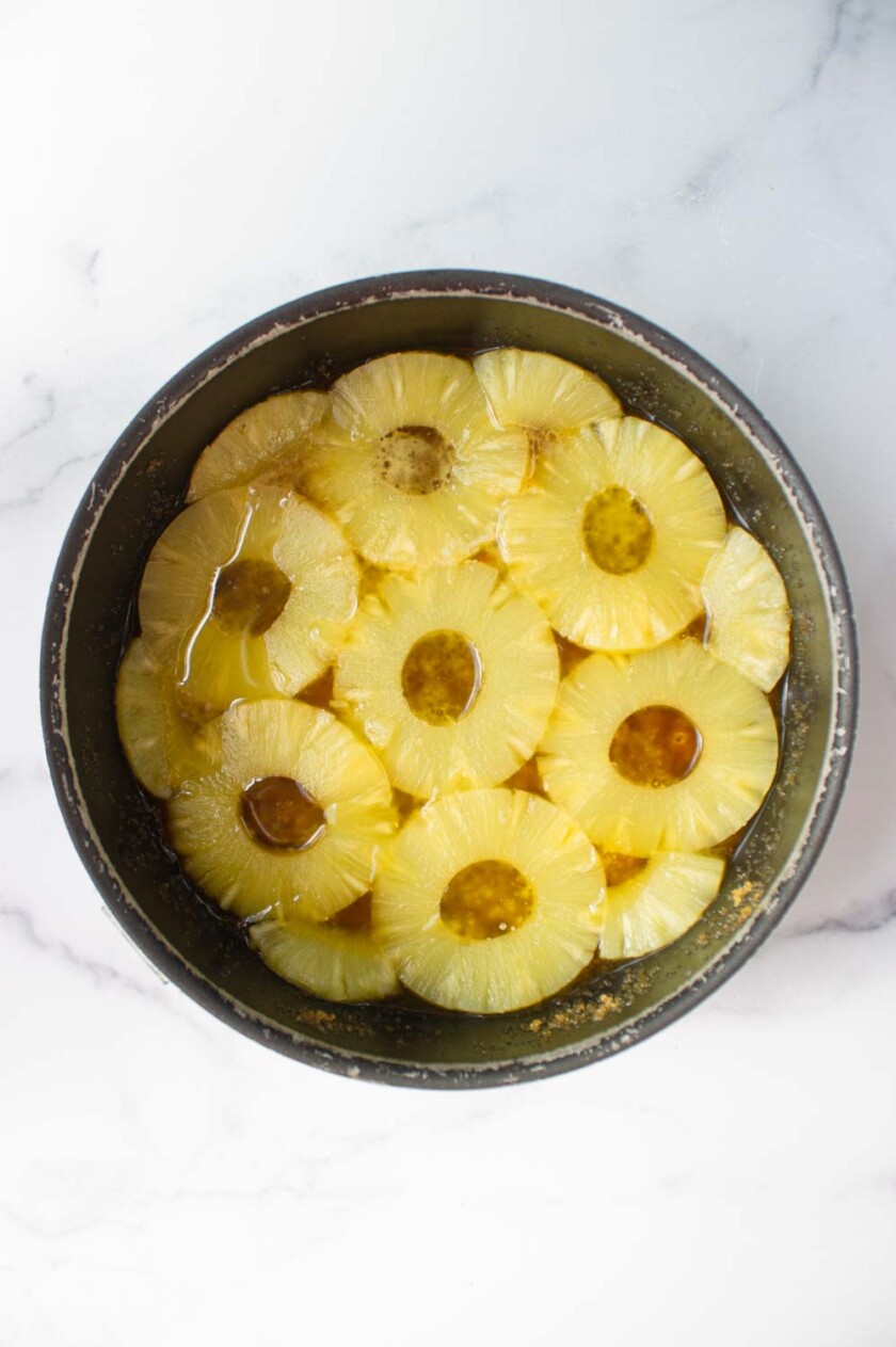 Torta-de-piña-volteada-by-enrilemoine-3 Una vista superior de un molde para hornear redondo conrodajas de piña cuidadosamente dispuestas con almíbar.