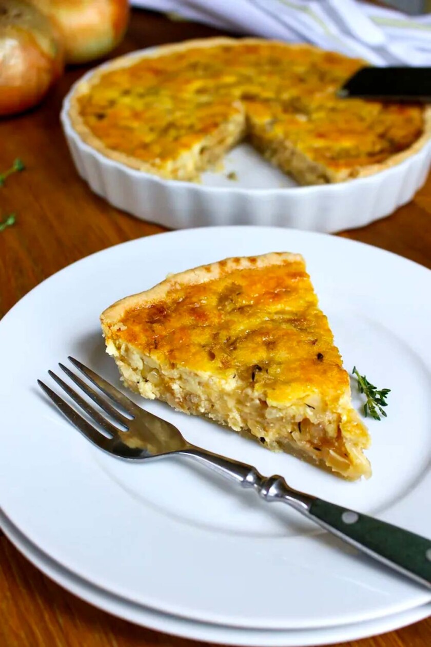 Una rebanada de quiche de cebolla en un plato blanco con un tenedor al lado, y el resto de la quiche en un molde para pastel al fondo.