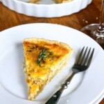 Una rebanada de quiche adornada con tomillo fresco en un plato blanco con un tenedor al lado. El resto de la quiche se ve en un plato blanco al fondo y cerca hay un vaso con una bebida de color claro.