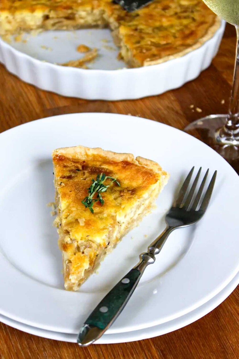 Una rebanada de quiche adornada con tomillo fresco en un plato blanco con un tenedor al lado. El resto de la quiche se ve en un plato blanco al fondo y cerca hay un vaso con una bebida de color claro.