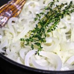 Un primer plano de cebollas en rodajas cocidas en una sartén con ramitas de tomillo encima.