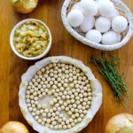 Una mesa de madera con cebollas, un plato de huevos, tomillo fresco, un plato de cebollas cocidas y una masa de tarta forrada con papel pergamino y rellena con pesas para tarta.