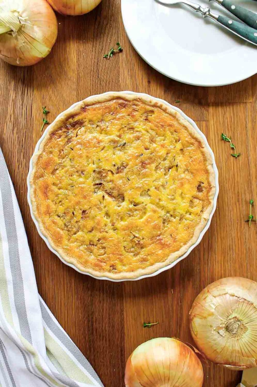 Quiche de cebolla Vidalia en un molde para tarta, colocada sobre una mesa de madera rodeada de cebollas enteras y un paño rayado. Sobre la mesa hay un plato blanco cercano con cuchillo y tenedor.