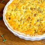Una quiche recién horneada en un plato de cerámica blanca se asienta sobre una superficie de madera cerca de una ramita de tomillo y una cebolla.