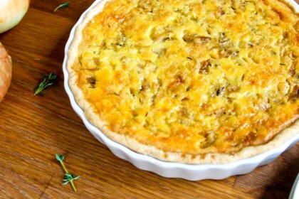 Una quiche recién horneada en un plato de cerámica blanca se asienta sobre una superficie de madera cerca de una ramita de tomillo y una cebolla.