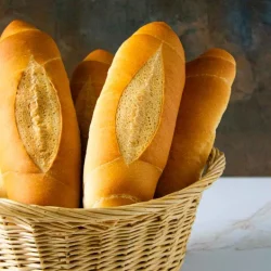 Una canasta de mimbre que contiene seis baguettes frescas de color marrón dorado, expuestas en posición vertical sobre un fondo oscuro con textura.