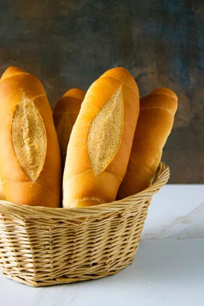 Una canasta de mimbre que contiene seis baguettes frescas de color marrón dorado, expuestas en posición vertical sobre un fondo oscuro con textura.