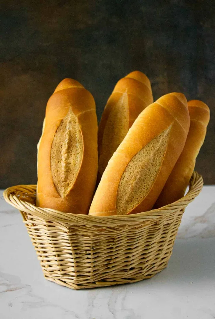 Una cesta de mimbre que contiene cinco baguettes se coloca sobre una superficie de mármol sobre un fondo oscuro.
