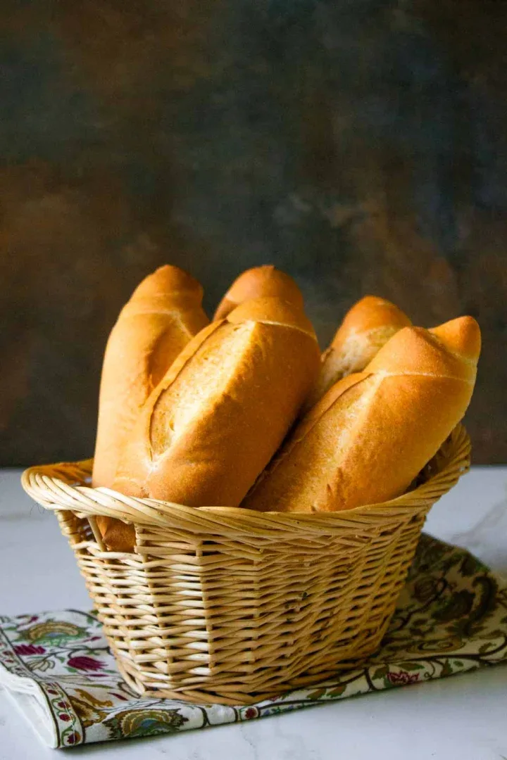 Una cesta de mimbre que contiene cuatro baguettes se encuentra sobre una superficie con una tela estampada debajo. El fondo es oscuro y borroso.