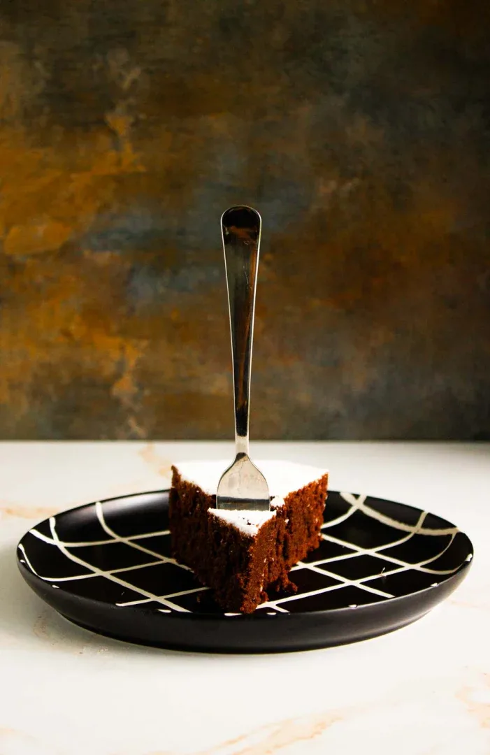 Un trozo de torta caprese de chocolate sobre un plato negro con líneas blancas, un tenedor en posición vertical en medio del pastel.
