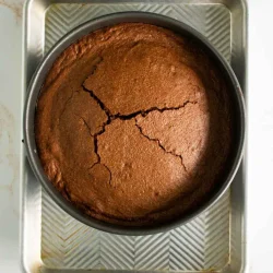 Un pastel de chocolate redondo con una superficie agrietada en un molde para hornear circular colocado sobre una bandeja de metal rectangular.