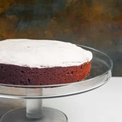 Una torta caprese de chocolate redondo espolvoreado con azúcar en polvo se asienta sobre un soporte de pastel de vidrio transparente sobre un fondo oscuro y texturizado.