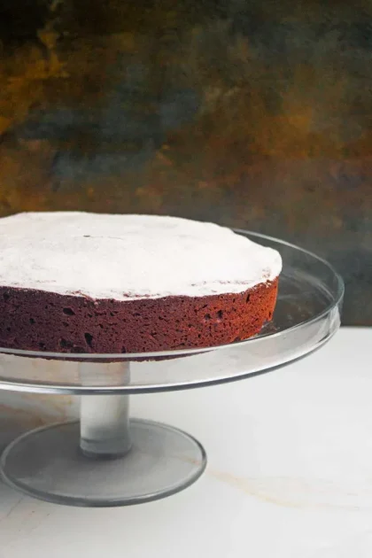Una torta caprese de chocolate redondo espolvoreado con azúcar en polvo se asienta sobre un soporte de pastel de vidrio transparente sobre un fondo oscuro y texturizado.