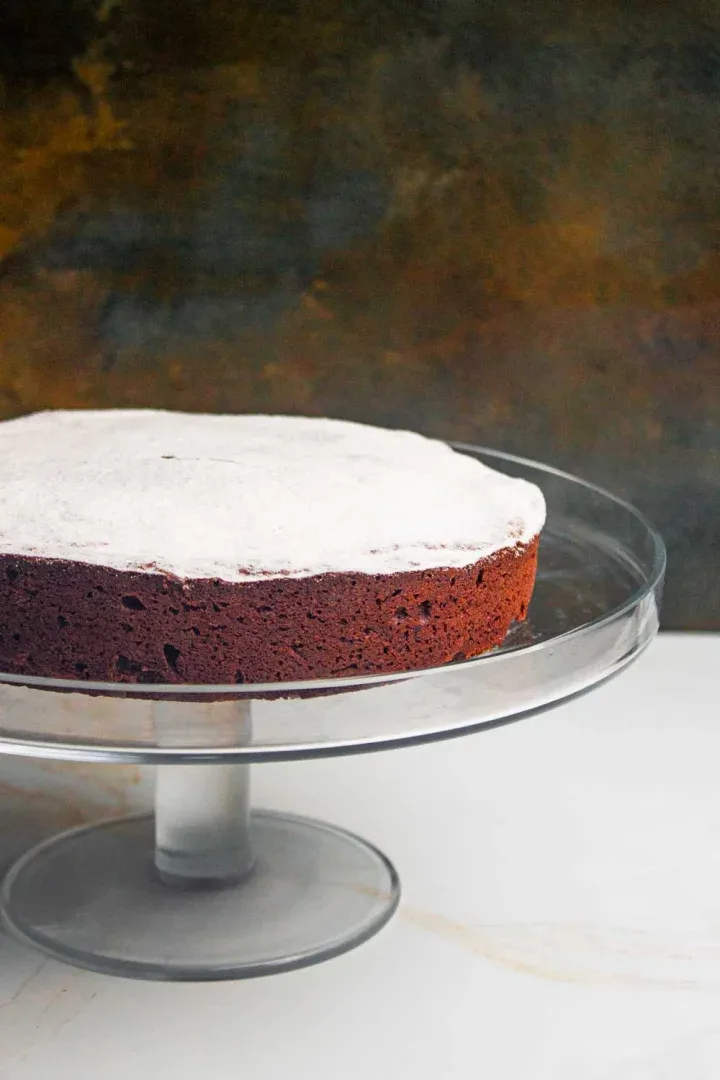 Una torta caprese de chocolate redondo espolvoreado con azúcar en polvo se asienta sobre un soporte de pastel de vidrio transparente sobre un fondo oscuro y texturizado.