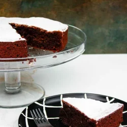 Una torta caprese de chocolate espolvoreado con azúcar en polvo se encuentra sobre un soporte de vidrio para pasteles, con una rebanada sobre un plato blanco y negro al lado de un tenedor.