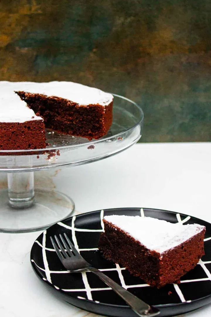 Una torta caprese de chocolate espolvoreado con azúcar en polvo se encuentra sobre un soporte de vidrio para pasteles, con una rebanada sobre un plato blanco y negro al lado de un tenedor.