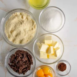 En recipientes separados se colocan varios ingredientes para hornear una torta caprese, incluidas claras de huevo, harina de almendras, azúcar, cubitos de mantequilla, chocolate picado, yemas de huevo, café en polvo y extracto de vainilla.