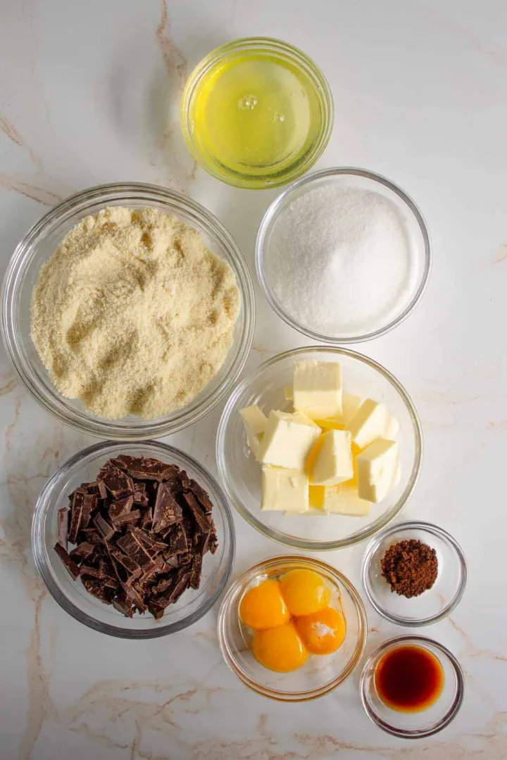 En recipientes separados se colocan varios ingredientes para hornear una torta caprese, incluidas claras de huevo, harina de almendras, azúcar, cubitos de mantequilla, chocolate picado, yemas de huevo, café en polvo y extracto de vainilla.