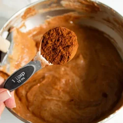 Una persona sosteniendo una cucharada de cacao en polvo sobre un tazón lleno de masa de chocolate.