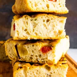 Una pila de cuatro porciones de focaccia con tomates cherry y romero sobre una superficie de madera.