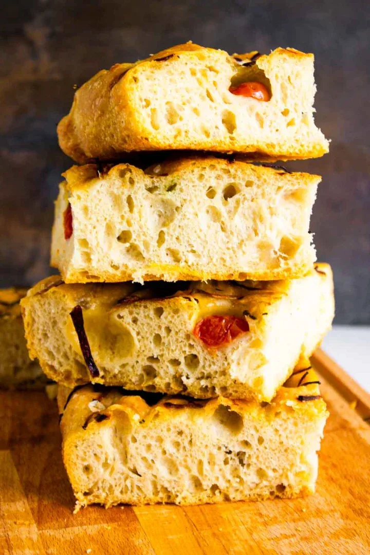 Una pila de cuatro porciones de focaccia con tomates cherry y romero sobre una superficie de madera.