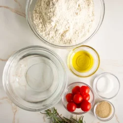 Una vista superior de los ingredientes para hornear pan focaccia: un recipiente de vidrio con harina, y tazones con agua, aceite de oliva, tomates cherry, sal, levadura y romero fresco sobre una superficie blanca.