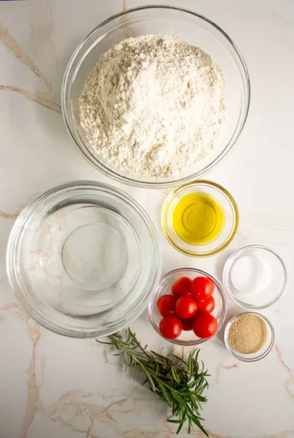 Una vista superior de los ingredientes para hornear pan focaccia: un recipiente de vidrio con harina, y tazones con agua, aceite de oliva, tomates cherry, sal, levadura y romero fresco sobre una superficie blanca.
