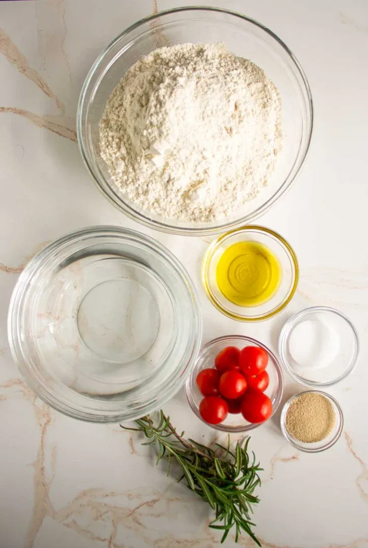Una vista superior de los ingredientes para hornear pan focaccia: un recipiente de vidrio con harina, y tazones con agua, aceite de oliva, tomates cherry, sal, levadura y romero fresco sobre una superficie blanca.