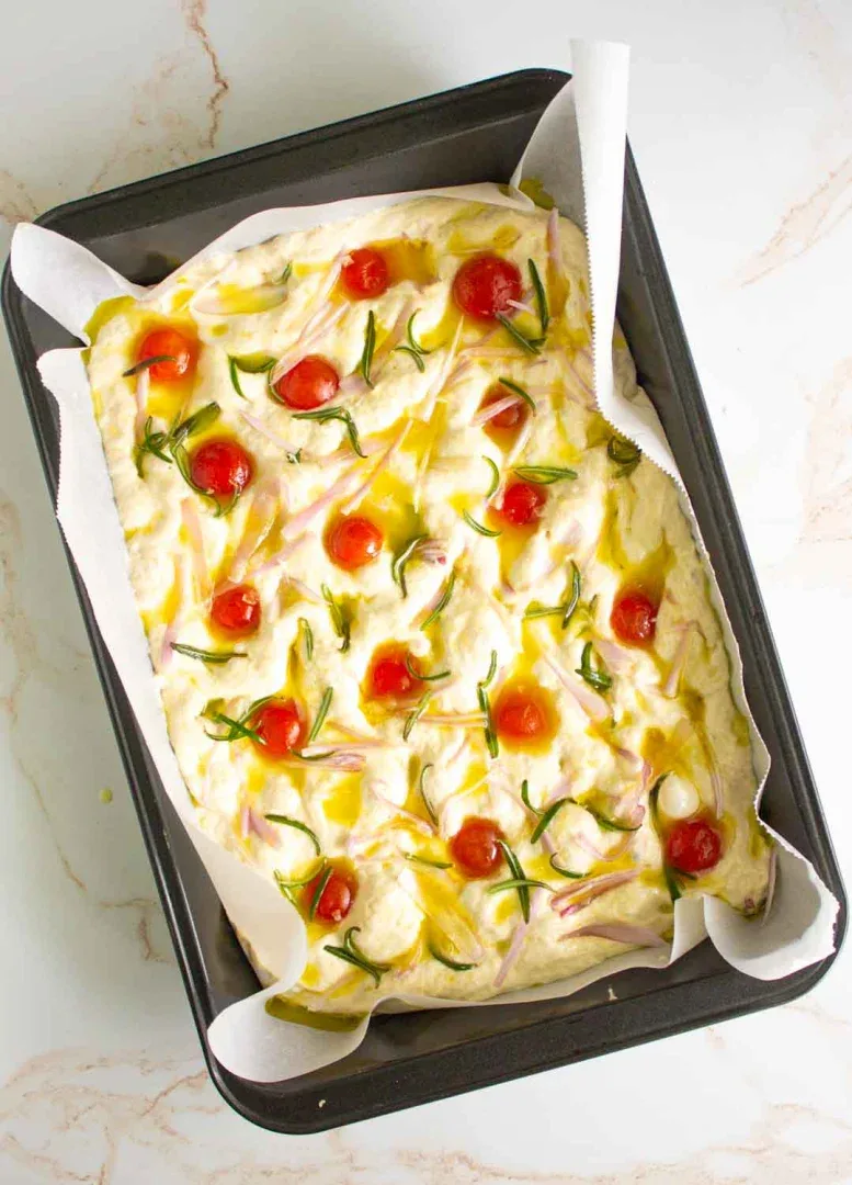Un molde rectangular con masa de focaccia cruda cubierta con tomates cherry, hierbas y aceite de oliva sobre papel pergamino.