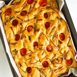 Focaccia rectangular cubierto con tomates cherry, romero y rodajas de cebolla en una bandeja para hornear forrada.