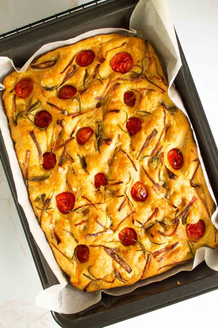 Focaccia rectangular cubierto con tomates cherry, romero y rodajas de cebolla en una bandeja para hornear forrada.