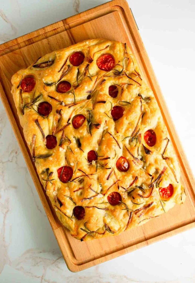 Focaccia rectangular cubierto con tomates cherry, romero y cebollas caramelizadas sobre una tabla de cortar de madera.