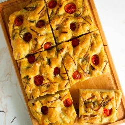 Focaccia rectangular cubierto con tomates cherry y hierbas se corta en trozos cuadrados sobre una tabla de cortar de madera.