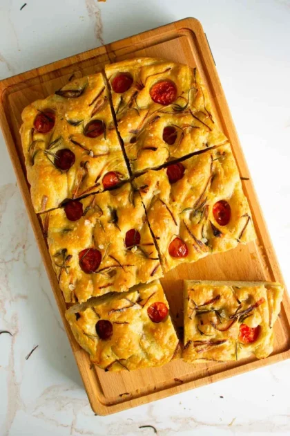 Focaccia rectangular cubierto con tomates cherry y hierbas se corta en trozos cuadrados sobre una tabla de cortar de madera.