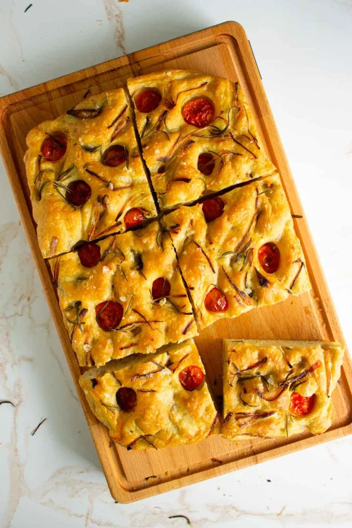 Focaccia rectangular cubierto con tomates cherry y hierbas se corta en trozos cuadrados sobre una tabla de cortar de madera.