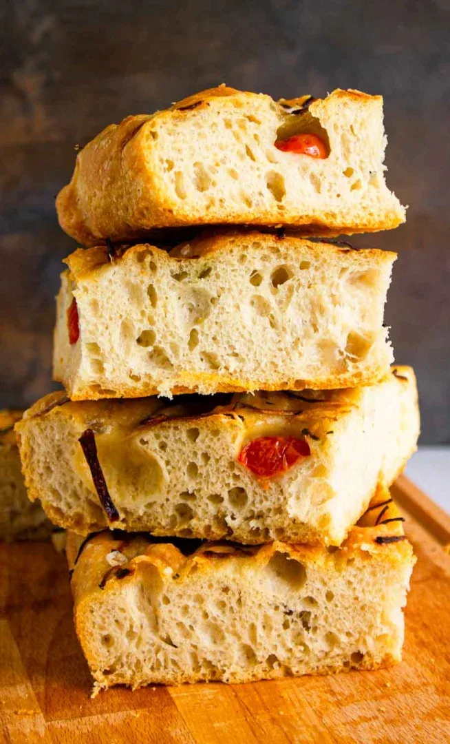 Una pila de cuatro rebanadas de focaccia con bolsas de aire visibles, tomates cherry y hierbas, colocadas sobre una superficie de madera.