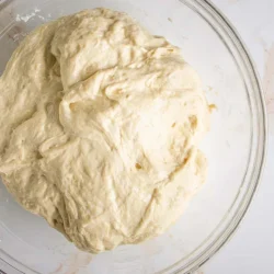 A ball of unbaked dough sits in a transparent glass bowl on a white marble surface.