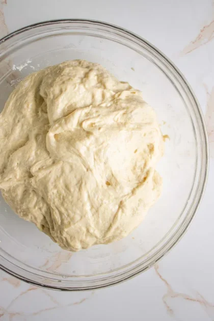 A ball of unbaked dough sits in a transparent glass bowl on a white marble surface.