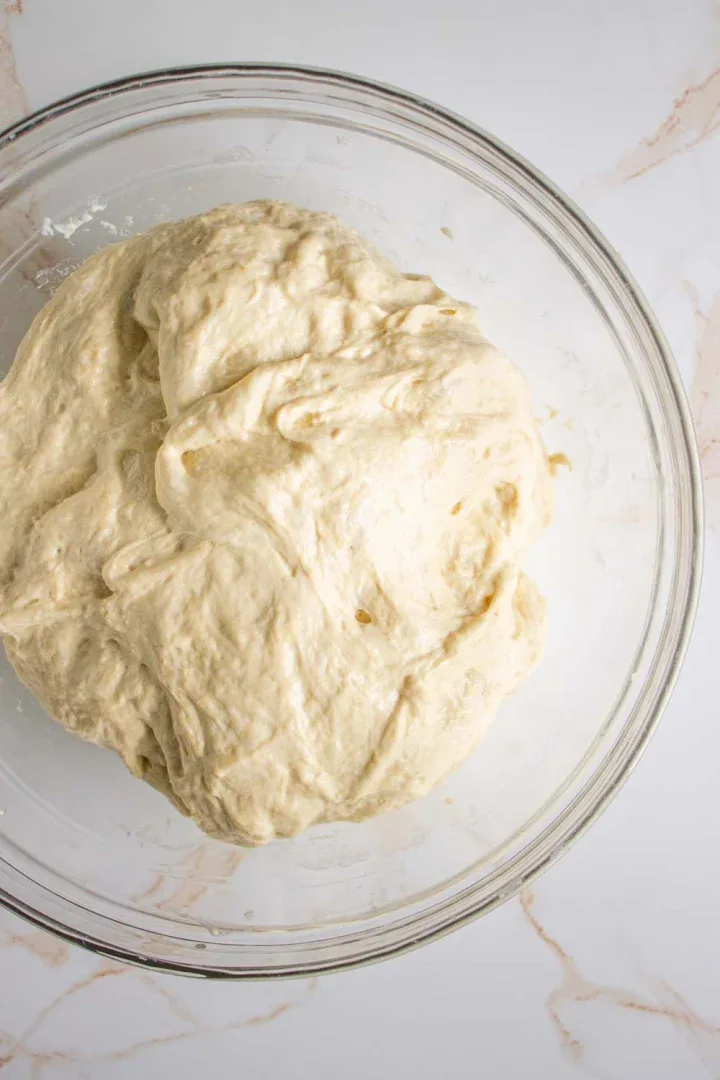 Una bola de masa cruda reposa en un recipiente de vidrio transparente sobre una superficie de mármol blanco.
