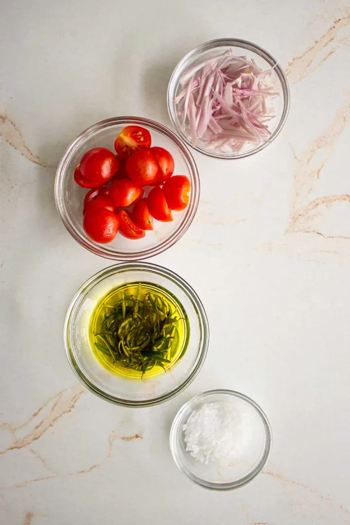 Cuatro cuencos sobre una superficie de mármol que contienen tomates cherry cortados por la mitad, chalotes en rodajas, aceite de oliva con romero fresco y sal gruesa.